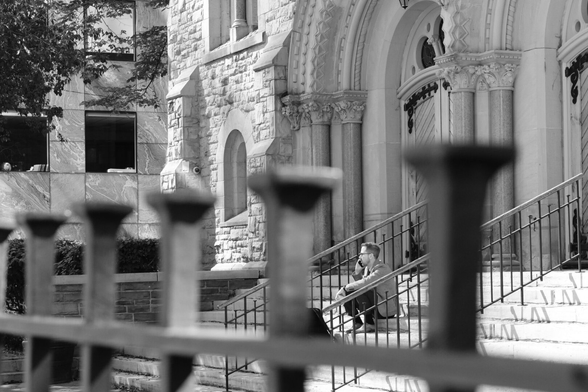 Outside a church. In the foreground is a slightly blurred wrought iron fence Framed by the fenceposts we can see a man sitting on the steps of the church, his phone to his ear. Behind him are the stone arches of the church’s doors.