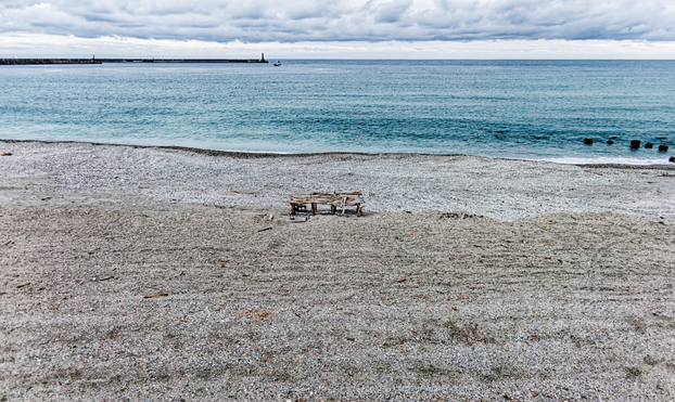 Pebble beach with with an unidentifiable decrepit wooden structure in the foreground, and the sea, a boat, and a jetty with a small lighthouse in the background.