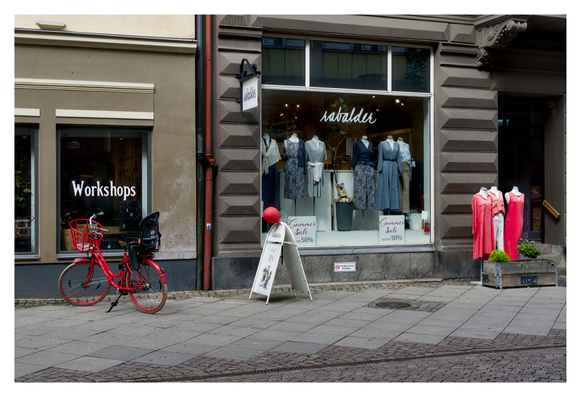 En liggande bild på ett par skyltfönster. Till vänster står en röd cykel, i mitten sitter en röd ballong och till höger hänger tre röda klänningar.