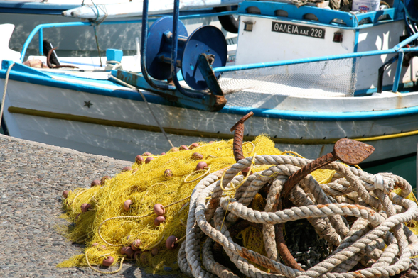 A colorful pile of fishing nets with floats, tangled ropes, and rusty anchors, situated on a stone dock beside a small blue and white fishing boat.
