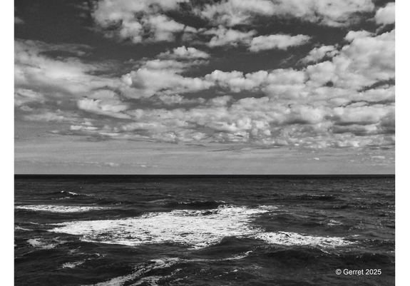 Black and white photo of an ocean with gentle waves under a dramatic, cloud-filled sky. The scene conveys a sense of calm and vastness.