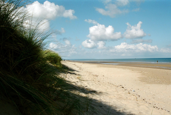Grassy dunes camera left, a sliver of sea camera right, a triangle of sandy beach mud frame, blue sky with fair weather clouds overhead.