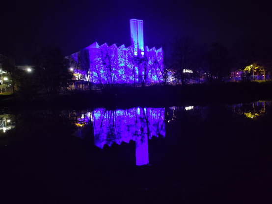 Das blau beleuchtete Gaskraftwerk spiegelt sich in der Saar. Vor dem Kraftwerk ist das Geäst von Bäumen ohne Laub.