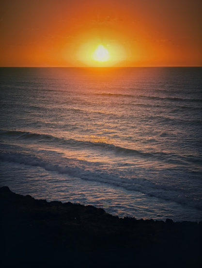 A vibrant vertical photograph of a dramatic sunset over the ocean. The sun, a bright orange orb, dominates the upper centre, casting an intense, fiery glow across the sky and reflecting on the water's surface. Below, the dark blue-grey waves of the sea are visible, with white foam breaking near the foreground. The bottom edge is dominated by a deep, dark silhouette of coastal land or cliff edge. The overall mood is powerful and tranquil.
