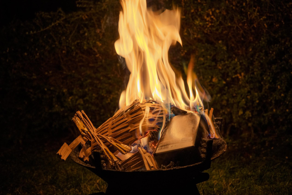 Night-time photo of a mini garden barbecue with cardboard boxes and other paper trash brightly burning inside. The tall intense bright flame rises up and lights up the surrounding garden in a warm, cozy glow.