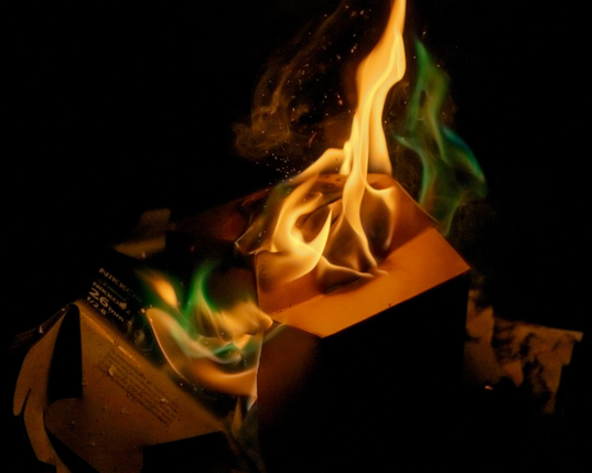 Night time photo of a bonfire where various boxes and other paper trash are being burned, notably the box of the lens on which the photo was taken. Alongside the main orange flame, there is an intense green flame that somewhat resembles an Aurora or Nebula.