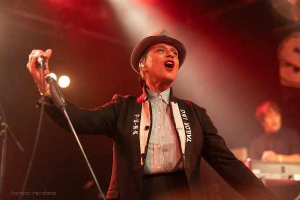 A performer passionately singing on stage, wearing a stylish outfit that includes a hat and a tailored jacket. The background features blurred lights and another musician playing an instrument.