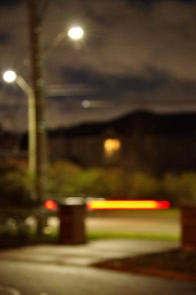 Blurry residential street scene:
A car’s light streaking by in red and yellow. Brick posts and a path on one side, bushes in-front of homes—one with a yellow light from a window—on the far side. Under a dark and light cloudy night sky on both sides, stand two lampposts with white lights.