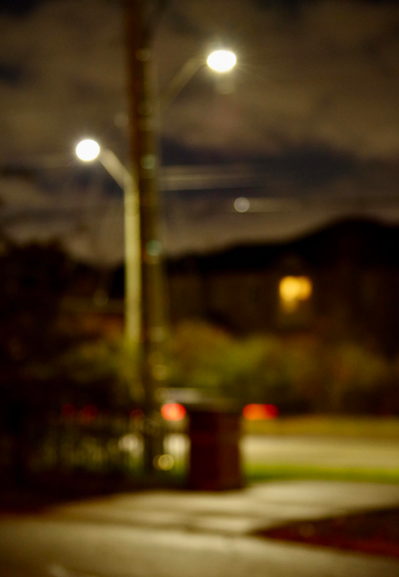 Blurry residential street scene:
A car’s light streaking by in red and yellow. Brick posts and a path on one side, bushes in-front of homes—one with a yellow light from a window—on the far side. Under a dark and light cloudy night sky on both sides, stand two lampposts with white lights.