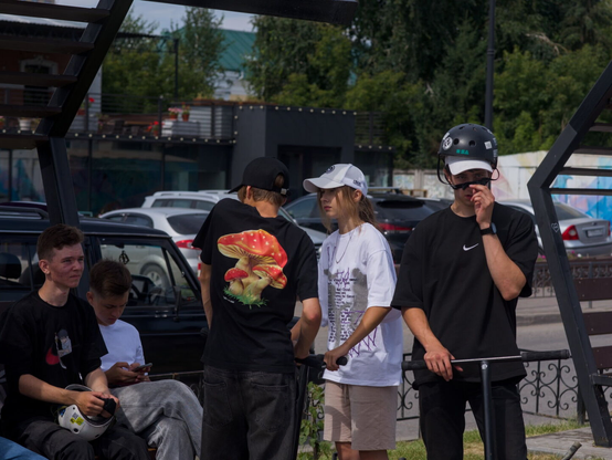 The image captures a group of teenagers gathered in an outdoor setting, possibly during the day given the natural light. The central focus is on five people standing and sitting around each other.

Starting from the leftmost side, there's a boy wearing black t-shirt and black jeans, seated on what appears to be a bench, holding onto a white helmet with both hands. Next to him seats another boy in white t-shirt and gray pants, holding what could be a phone. Next to them stands another person wearing a black t-shirt adorned with vibrant red and yellow graphics, possibly depicting mushrooms. Next there is a girl dressed casually in a white t-shirt, beige shorts and white cap. On the right side, there is an boy, he's adjusting his sunglasses given, and he's also is wearing a helmet for protection. The helmets worn by some individuals hint at recent participation in some sports activities.

The backdrop of this scene includes various parked cars and trees.