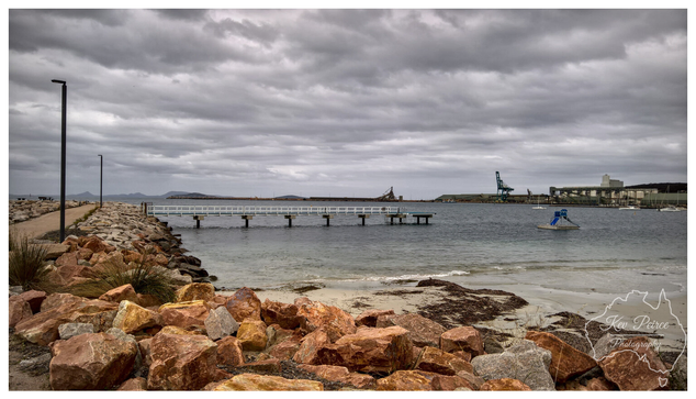 A wide angle landscape photograph of the Esperance Tanker Jetty area on a cloudy, overcast day.  In the foreground, a pile of large, reddish-orange granite boulders forms a natural barrier leading up to a paved walking path on the left.  The water is calm, meeting a small sandy beach patch with seaweed. A long, low white jetty extends into the water toward the center.  In the background, industrial elements of the working port are visible, including a large green crane and port buildings, backed by distant hills under a vast, gray sky.