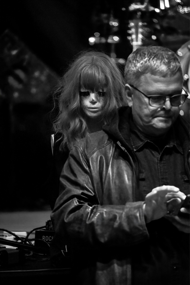 Ein Mann steht mit dem Rücken zu einer Bühne und schaut in sein Handy. Hinter ihm, auf der Bühne, steh der von ihm mitgebrachte Torso einer weiblich gelesenen Schaufensterpuppe. Ihr Kopf  schaut über seine rechte Schulter.