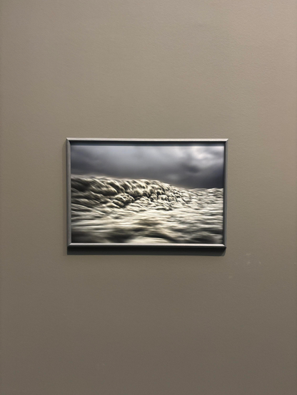 A framed photograph of waves, displayed on a neutral-colored wall. The image features a monochromatic palette with cloudy skies above.