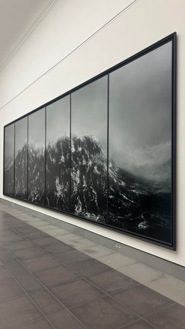 A large black-and-white photograph of a powerful ocean wave is displayed on a wall. The artwork is divided into several panels, emphasizing the movement and depth of the water. The exhibition space features neutral-colored flooring and walls, enhancing the visual impact of the scene