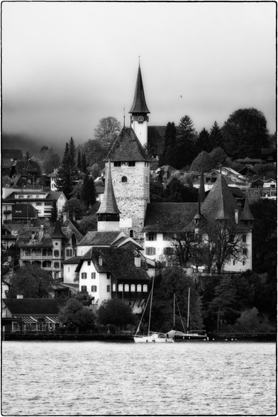 Eine Schwarz-Weiss-Aufnahme zeigt eine malerische Dorfszene an einem Seeufer.

Im Zentrum steht eine markante mittelalterliche Struktur mit einem hohen, steinernen Turm und einem spitz zulaufenden Dach. Oberhalb davon ist eine höhere Kirche mit einem schlanken Turm und einer Uhr sichtbar. Die Gebäude sind dicht in den Hang eingebettet, umgeben von Bäumen und Wohnhäusern, die sich terrassenförmig erstrecken.

Im Vordergrund liegt eine spiegelglatte Wasserfläche, auf der zwei Segelboote vertäut sind. Die Szene ist von einer nebligen oder bewölkten Atmosphäre überzogen, was den Kontrast und die dramatische Wirkung der Schwarz-Weiss-Aufnahme verstärkt.