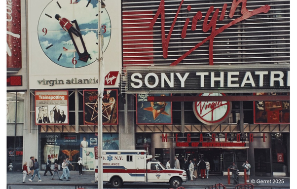 Street scene in front of a Virgin-branded Sony Theatre. Large Virgin Atlantic clock and movie posters visible. Busy sidewalk with pedestrians and NY ambulance.