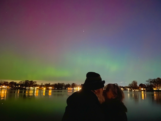 Photograph of a couple kissing in front of a bright green and red aurora over a lake.