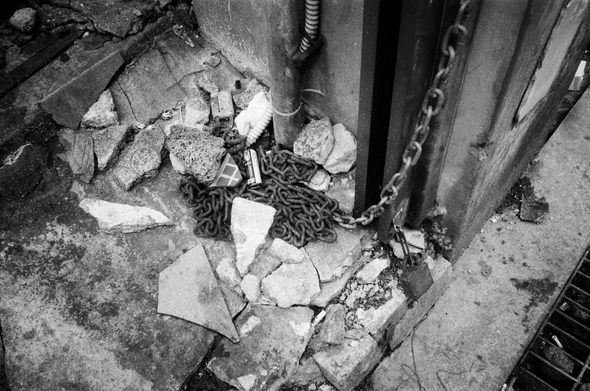 주택가 벽면에 매달린 쇠사슬이 또아리를 튼 것처럼 바닥에 깔려있고, 그 위로 깨진 콘크리트 덩어리가 몇 개 떨어져있다. 주변에는 에어컨 관, 자물쇠, 하수구 등이 보인다.