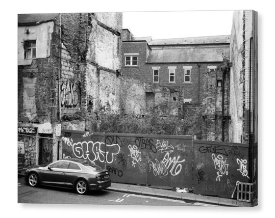 Black and white photo of a derelict plot between 2 graffiti covered brick buildings.  The image is shown printed upon a block canvas.