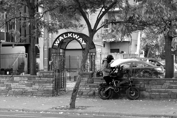 In the centre of the image, a man in a helmet stands beside a bike, looking in his bag. To his left is a wrought iron arch with a sign that reads “Walkway” Between them is a slender, sinuously curving tree. Behind them are the trappings of the city: an apartment building, a parking lot, and more trees.