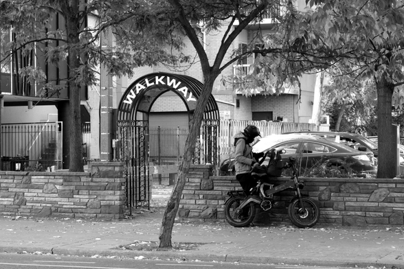 In the centre of the image, a man in a helmet stands beside a bike, looking in his bag. To his left is a wrought iron arch with a sign that reads “Walkway” Between them is a slender, sinuously curving tree. Behind them are the trappings of the city: an apartment building, a parking lot, and more trees. 