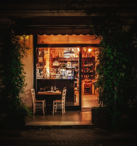 This is a photo of a cozy, warmly lit restaurant at night. The entrance is framed by green bushes. Outside the entrance on the left there is a wooden table with two white chairs which sit near the glass door. Inside through the windows there are shelves filled with glassware and bottles line the back wall. The photo is centrally lit with soft, yellow overhead lights.