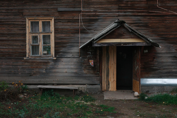 The photo shows the entrance to an old wooden house. There is a triangular canopy over the entrance. The door is open. There is a window and a wooden bench next to the entrance. The front of the house has green grass.