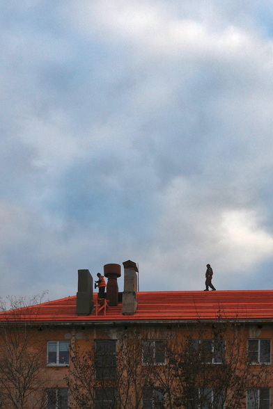 Workers are repairing the red roof of a five-story building. The sky above the roof is covered with clouds.