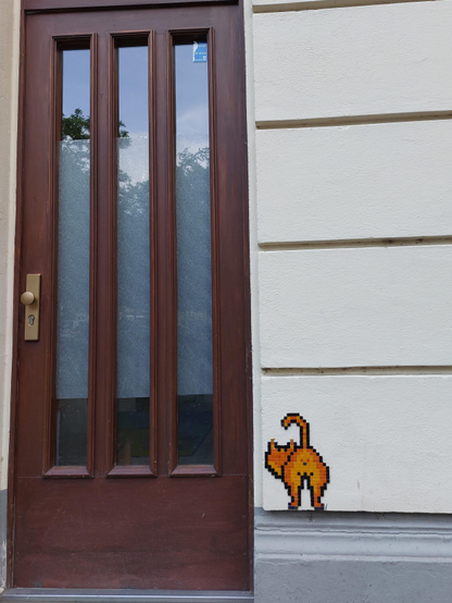 A simple brown wooden front door with three vertical glass inserts. Trees and a patch of blue sky are reflected in the glass. To the right of the door, the façade is structured by horizontal white stucco panels. A small mosaic in shades of orange is attached to the bottom panel. It shows a cat from behind with its tail stretched out adventurously, its head turned to the left.
A small, cheerful respite from the hustle and bustle of the city!
