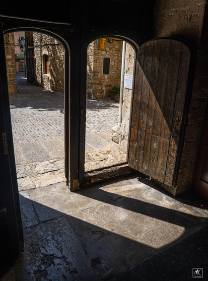Color photo taken from inside the shadowed entry space of a church looking out two adjacent, open, arch-topped doors at a sunlit cobblestone street beyond.