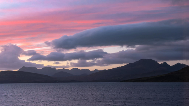Sunset over the Cuillin mountains with wispy red colouration sitting on top of the usual grey cloud with the jagged silhouette of the mountain range underneath. 