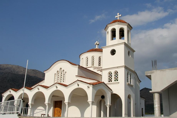 A white church with a red-tiled roof, featuring a bell tower and several arched entrances. The structure is set against a mountainous backdrop under a partly cloudy sky.