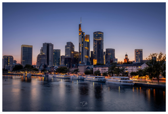 View of the skyline of Frankfurt with a river and boats in front photographed from a bridge. The photo was taken right after sunset, so the skyline and the waterfront path are illuminated.