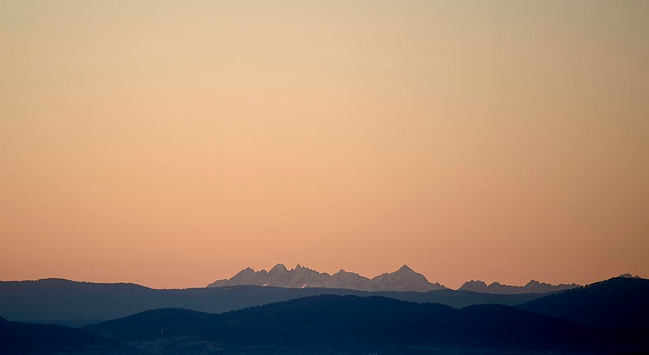 This photo shows a tranquil mountain landscape at sunrise. In the foreground are dark, gently rolling hills. Behind these, a shadowy, higher ridge stretches horizontally across the frame. In the distance, a line of dramatic, jagged peaks is softly illuminated by the first warm, orange-pink light of dawn. Above these mountains, the clear sky glows with gentle pastel tones, filling the scene with a calm, peaceful atmosphere. There are no visible human figures, buildings or vegetation, just natural silhouettes and the quiet beauty of the sunrise over the mountains.