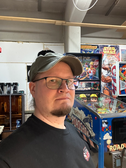 A selfie-style picture of a Nordic looking guy in his mid-40s. I'm wearing a green, worn baseball cap, glasses and a short beard. The background shows a workshop, with a 3D printer and couple of work in progress pinball machines.