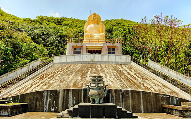 Top of a temple with an incense burner in the foreground and a large golden bodhisattva statue against green hills in the background.