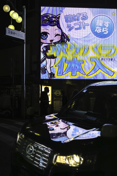 A man is walking by an illuminated door in a dark little street in Tokyo at night, his little black silhouette is illuminated in a yellow glow. Hanging above him, a big flashy neon advertisement sign with a manga character, making him look even smaller, is reflected on a black car passing by in the foreground. On the top left corner, an art deco street lamp and street sign of the Kabukicho neighbourhoud of Tokyo adds another touch of light and color in this dark scene.  