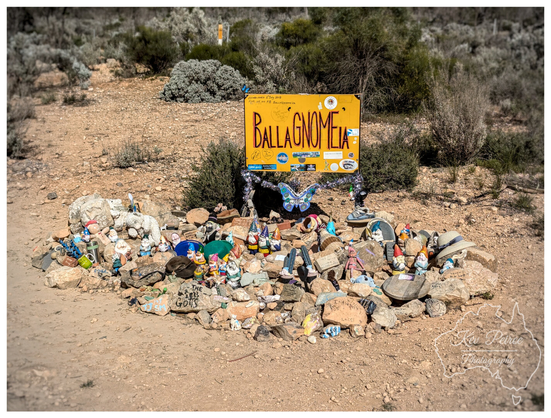 A wide angle photo of a yellow wooden sign in a dry, scrubby roadside environment. The sign, hanging between two posts wrapped in fairy lights, reads "BALLAGNOMEIA" in large orange, whimsical text.

Below the sign is a large cairn or pile of rocks covered in dozens of small garden gnomes and various trinkets, including a blue butterfly decoration, a small toy goat, and some hats. The ground is dry dirt and gravel.