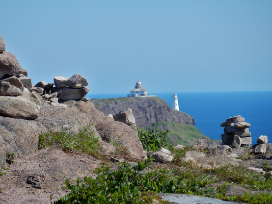 In the foreground is a rocky landscape with a large body of water in the background. There are also some stacked rocks on either side of the foreground frame. Also in the distance is lighthouse with a building next to it, however the perspective makes it look like the lighthouse and building are the same distance as the rocks and are just very, very tiny.