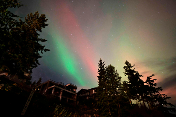 Aurora against a hazy sky. One bright red band and one brighter green band, with some other red and green to the side. They are framed by tall evergreens and "small" mansions.
