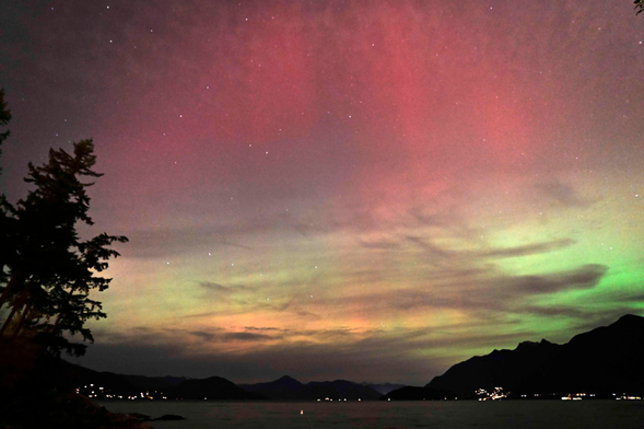 Red and green aurora in a cloudy sky, with mountains, distant lights, and the sea in the foreground.
