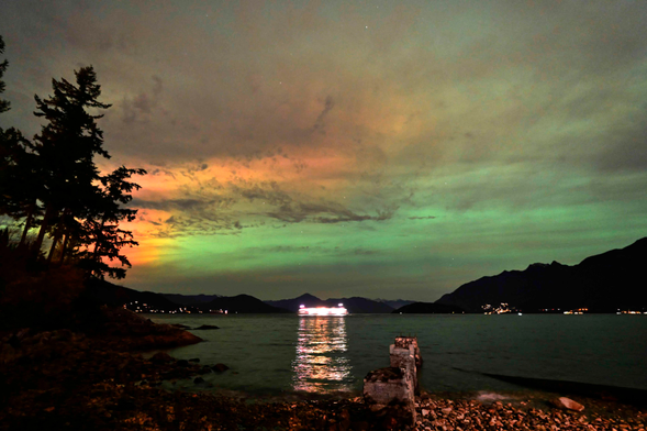 Green aurora, with a bit of red, in a mostly clouded sky. In the centre of the frame, a large ferry passes through, its lights reflecting across the sea right to a rocky beach.