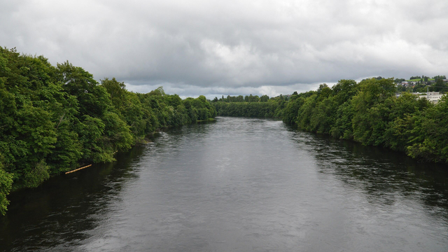 A photo of a river with trees lining both sides. The sky is filled with clouds.