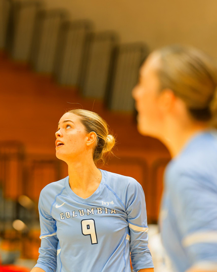 Blonde woman with her hair tied into a single bun in back is looking up and wearing a light blue shirt that says COLUMBA 9 while another closer women is looking the same way but too close to be in focus,  and the empty bleachers behind are too far to be in focus