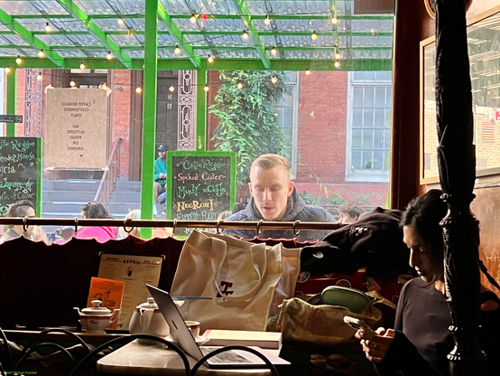 An image - photo by Fenichel - from within  historic  Café Reggio in the heart of NYC's "Greenwich Village"/. Backlit in the background (upper half) shows a sunny MacDougal Street, with brownstones and red brick buildings with steps, as well as a big potted plant. But in the center is someone outside, who might be looking in, but more likely looking at his phone, like the person "on the inside" (lower right) silhouetted in the light outside. Center bottom is an open computer, tea pot and shopping bag. Separating inside and outside is a brass curtain rod with curtains below.
