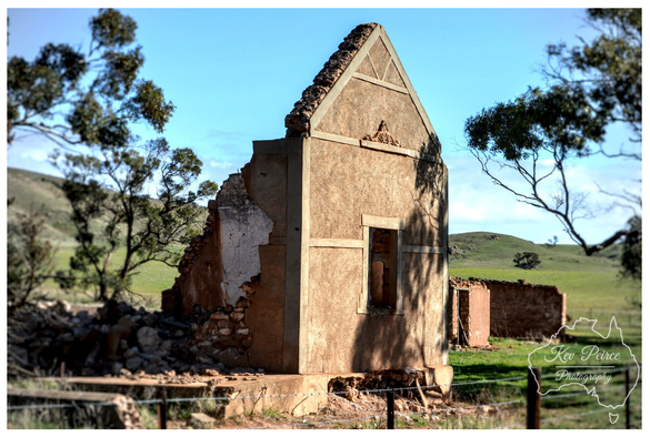 The sunlit, crumbling stone facade of an old, abandoned building ruin in Eudunda, South Australia.  The central gable structure has an empty window opening, and the walls are partially fallen away, revealing rubble and internal stones.  It is surrounded by green rolling hills, scattered eucalyptus trees, and a barbed wire fence in the foreground.  Photo by Kev Peirce.