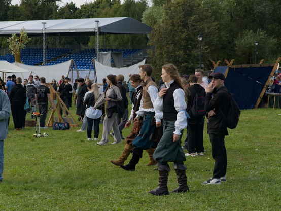The image shows a grassy outdoor area, likely a park or field, filled with people. In the background, there’s a large white canopy or tent structure and a blue seating area with a blue railing. 

A group of several people are prominently featured in the foreground. They are dressed in historical clothing – men wearing brown leather vests over white shirts, dark brown trousers, and black boots. The women are wearing similar attire, with longer hair. They are walking in a line towards the left side of the frame.

Behind the walking group is a temporary structure – a white tent or canopy with a blue railing. In the distance, there's a seated area with blue plastic chairs and a blue railing around it. Several other people are scattered throughout the background, some standing and watching.

The scene is illuminated by natural daylight. The dominant colors are green (grass), brown (clothing), white (tent), blue (seating), and the clothing shades of the people.