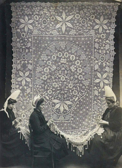 vers 1920, des femmes bretonnes, dentellières à la main, en tenue et coiffe traditionnelle travaillent en commun sur une dentelle suspendue au mur