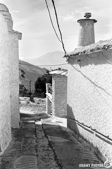 A narrow part cobbled street winds between whitewashed buildings with chimneys in Capileira, a mountain village, under clear skies. Distant mountains rise in the background. Black and white photo.