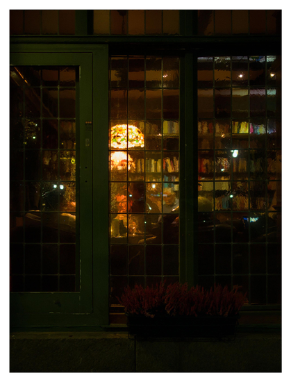 En stående bild på några fönster till en pub. Fönstren är gjorda så att det på andra sidan blir oklart. Två lampor i Tiffany-stil hänger från taket, människor samtalar och bokhyllor står i bakgrunden.